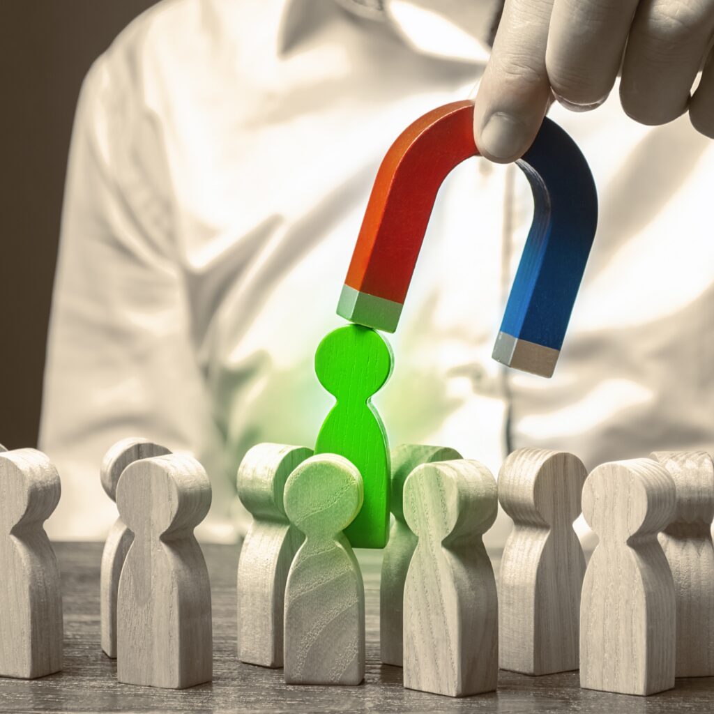 An adult's hand holds a magnet attracting a new customer over a crowd of wooden figures standing on a wooden surface.